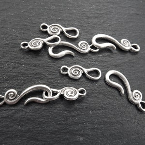 May include: A collection of antique silver-coloured jewellery clasps featuring a swirling design. The clasps vary in size and shape, with a hook and eye closure. These metal clasps are likely for necklaces or bracelets.