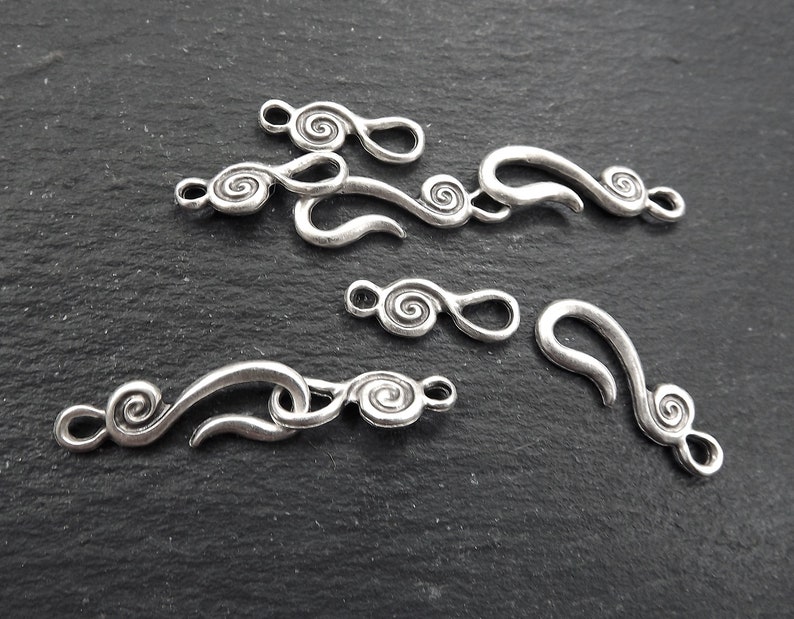 May include: A collection of antique silver-coloured jewellery clasps featuring a swirling design. The clasps vary in size and shape, with a hook and eye closure. These metal clasps are likely for necklaces or bracelets.