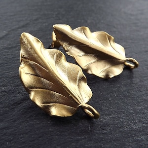 Puede incluir: Dos abalorios de hoja de oro con un bucle en la parte inferior. Las hojas tienen una superficie texturizada detallada.