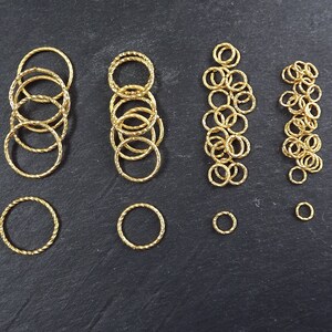 May include: Assortment of gold jump rings in various sizes, perfect for jewelry making. The rings are arranged in rows on a black surface.