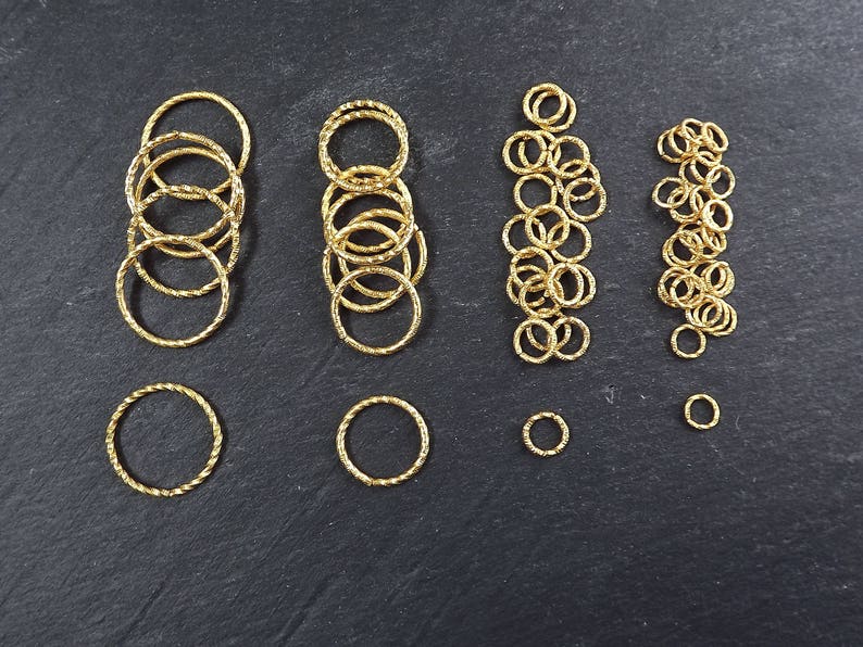 May include: Assortment of gold jump rings in various sizes, perfect for jewelry making. The rings are arranged in rows on a black surface.
