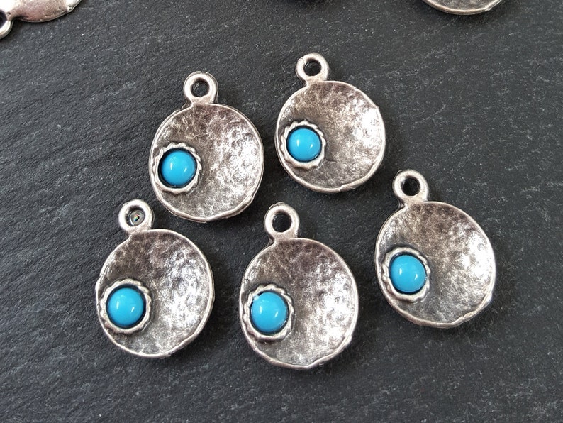 May include: Five silver-toned metal charms with a hammered texture and a turquoise blue cabochon stone in the center of each charm.