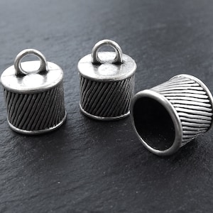 Könnte beinhalten: Drei silberne Metall-Kordelenden mit einem dekorativen, strukturierten Design. Jede Kappe hat eine kleine Schlaufe oben.