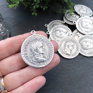 May include: Silver-toned metal charms featuring a Roman emperor's profile and inscription. The charms have a circular shape with a raised edge and a loop for attaching to jewelry or other crafts.