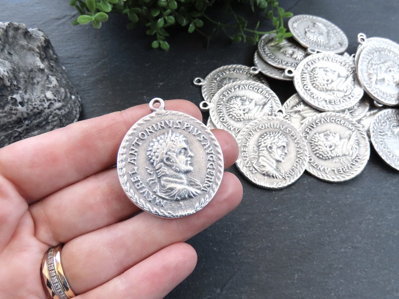 May include: Silver-toned metal charms featuring a Roman emperor's profile and inscription. The charms have a circular shape with a raised edge and a loop for attaching to jewelry or other crafts.