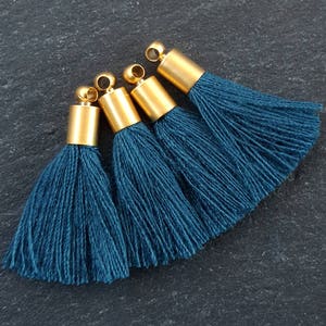 May include: Four teal blue tassel charms with gold metal caps. The tassels are made of soft, fluffy yarn and are perfect for adding a touch of color and texture to your crafts.