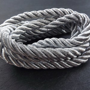 Puede incluir: Cordón trenzado plateado, una cuerda gruesa y retorcida con un brillo metálico. El cordón está enrollado en un círculo, mostrando su textura y brillo.