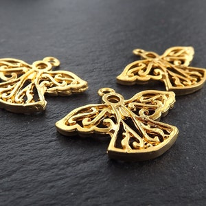 Puede incluir: Tres colgantes dorados con forma de &aacute;ngel, con intrincadas alas y cuerpos de filigrana. Cada colgante tiene un peque&ntilde;o lazo en la parte superior para sujetarlo a la joyer&iacute;a. Los colgantes se muestran sobre una superficie oscura y texturizada.