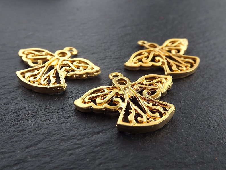 Puede incluir: Tres colgantes dorados con forma de &aacute;ngel, con intrincadas alas y cuerpos de filigrana. Cada colgante tiene un peque&ntilde;o lazo en la parte superior para sujetarlo a la joyer&iacute;a. Los colgantes se muestran sobre una superficie oscura y texturizada.