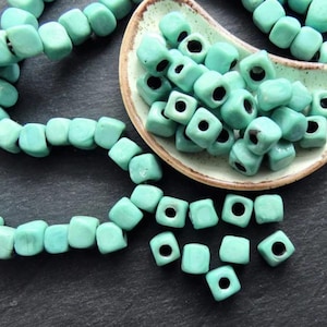 以下が含まれることがあります： ターコイズブルーのセラミック製の正方形のビーズのストランド。同じビーズが浅い皿に少量置かれている。