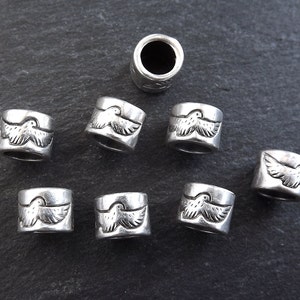 May include: Eight silver metal beads with a bird design. The beads are cylindrical and have a hole in the center. They are arranged in a circle on a gray surface.