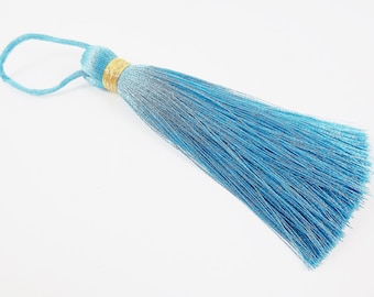Puede incluir: Un borla de color azul claro con ribete dorado. La borla está hecha de fibras suaves y sedosas y tiene un lazo en la parte superior para colgar.