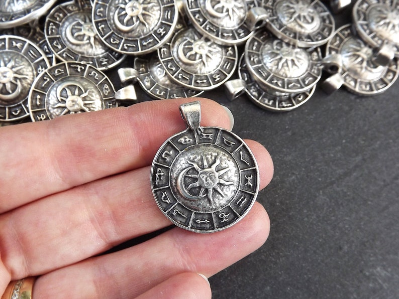 May include: Silver pendant with a circular design featuring a sun and moon symbol surrounded by zodiac signs.