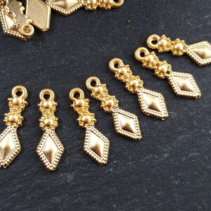 May include: Eight gold-toned metal pendant charms with a diamond shape and three small round beads at the top. The charms are arranged in a row on a dark gray surface.