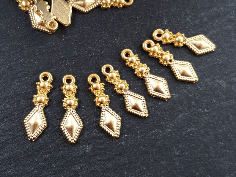 May include: Eight gold-toned metal pendant charms with a diamond shape and three small round beads at the top. The charms are arranged in a row on a dark gray surface.