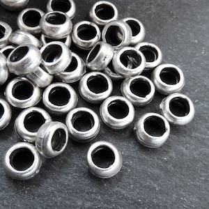 Op de afbeelding: Een stapel zilverkleurige metalen kralen met een ronde vorm en een gat in het midden. De kralen zijn willekeurig gerangschikt op een donkergrijs oppervlak.