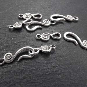 May include: Several silver-coloured metal clasps and connectors with a swirling, scroll-like design. The clasps vary in size and shape, with a hook-and-eye closure. The pieces are arranged on a dark, textured surface, suitable for jewellery making.