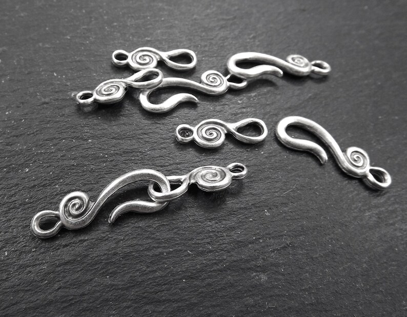 May include: Several silver-coloured metal clasps and connectors with a swirling, scroll-like design. The clasps vary in size and shape, with a hook-and-eye closure. The pieces are arranged on a dark, textured surface, suitable for jewellery making.