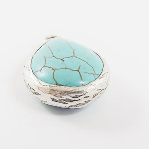 Op de afbeelding: Een druppelvormige hanger met een turquoise steen en een getextureerde zilveren rand. De steen heeft een gebarsten patroon. De hanger staat op een witte achtergrond. Sieraden.