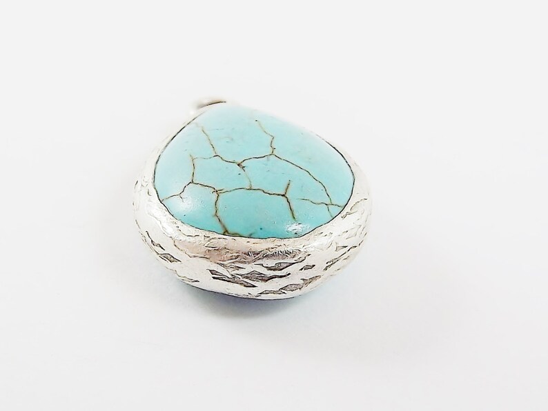 Op de afbeelding: Een druppelvormige hanger met een turquoise steen en een getextureerde zilveren rand. De steen heeft een gebarsten patroon. De hanger staat op een witte achtergrond. Sieraden.
