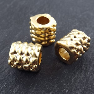Könnte beinhalten: Drei goldfarbene Metallperlen mit einem strukturierten, blumenartigen Design. Die Perlen haben ein Loch in der Mitte zum Auf fädeln.