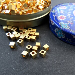 May include: A collection of small, gold-colored, square beads with a hole in the center. Some beads are scattered on a dark surface, while others are in a round, metal container. A blue, patterned container lid is also visible.