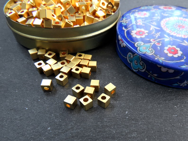 May include: A collection of small, gold-colored, square beads with a hole in the center. Some beads are scattered on a dark surface, while others are in a round, metal container. A blue, patterned container lid is also visible.
