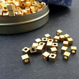 May include: A collection of small, square, gold-colored metal beads with a hole through the center. Some beads are scattered on a dark surface, while others are inside a round, metal tin.