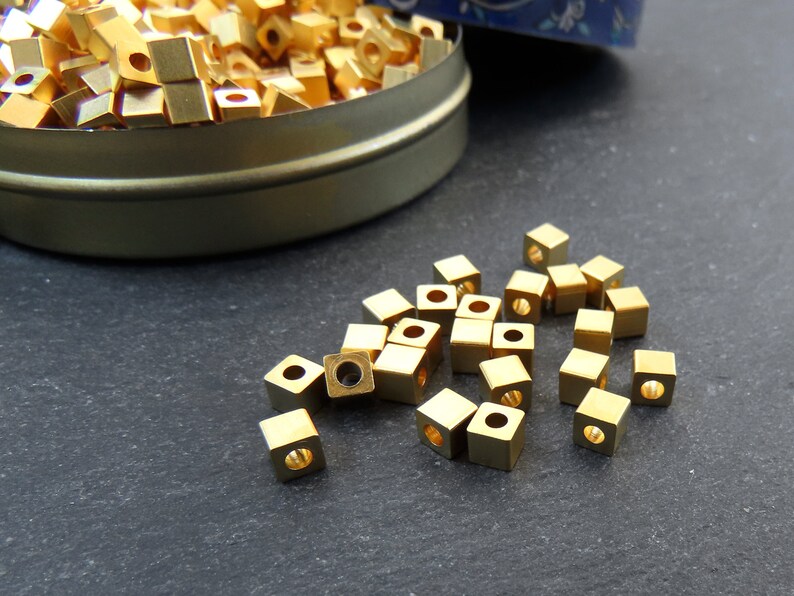 May include: A collection of small, square, gold-colored metal beads with a hole through the center. Some beads are scattered on a dark surface, while others are inside a round, metal tin.