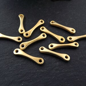May include: A group of 12 gold-colored metal connector charms with a curved shape. The charms have a hole at each end for attaching to jewelry or other crafts.