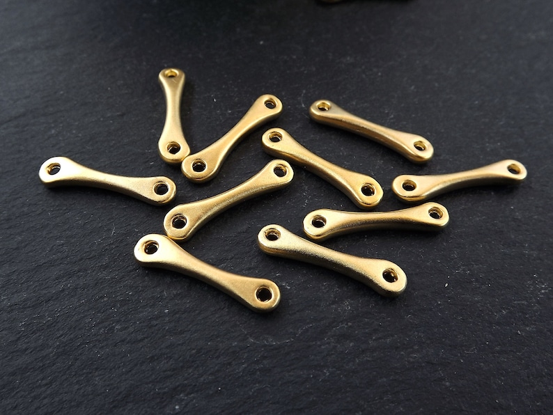 May include: A group of 12 gold-colored metal connector charms with a curved shape. The charms have a hole at each end for attaching to jewelry or other crafts.