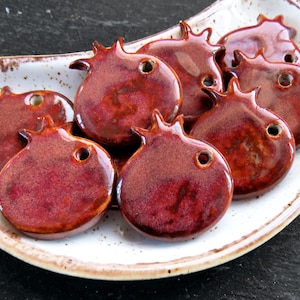 Può includere: Una collezione di ornamenti a forma di melograno in ceramica smaltata, nei toni del rosso e del marrone, disposti su un piatto bianco e marrone macchiettato. Ogni ornamento ha un piccolo foro e una superficie strutturata.