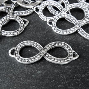 May include: A pile of silver infinity symbol charms with a beaded texture. The charms have a loop on one side for attaching to jewelry.
