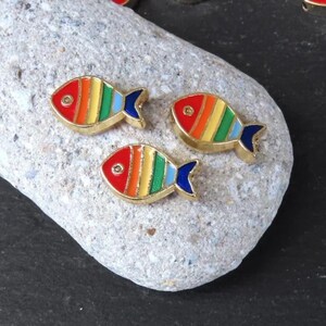 Puede incluir: Tres abalorios de pescado de color dorado con rayas de arcoíris y aletas azules. Los peces están sobre una roca gris.
