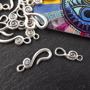 May include: Close-up of several silver metal hook and eye clasps. Each clasp has a decorative spiral design on both ends. The clasps are scattered on a dark surface, with a colourful patterned background visible.