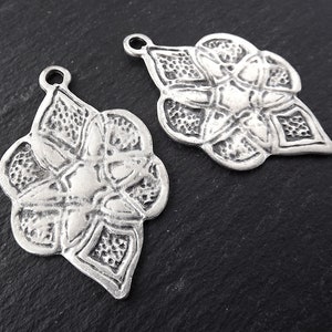 Könnte beinhalten: Zwei silberne Metall Anhänger in Form einer stilisierten Blume mit einem Loch oben zum Anbringen an einer Halskette oder einem Armband.