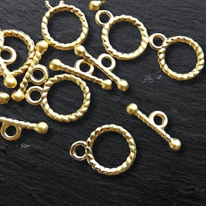 Puede incluir: Primer plano de cierres de palanca de metal dorado con un diseño de cuerda trenzada. Los cierres están esparcidos sobre una superficie negra.