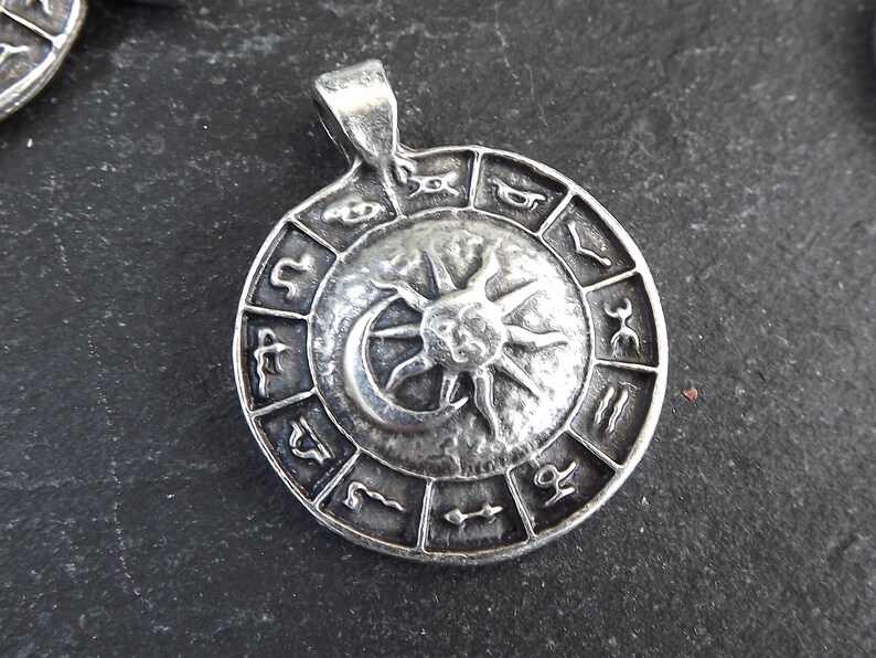 May include: Silver pendant featuring a circular design with a sun and moon in the center, surrounded by zodiac symbols.