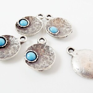 May include: Five silver charm pendants with a turquoise blue stone in the center. The pendants are round and have a textured surface.