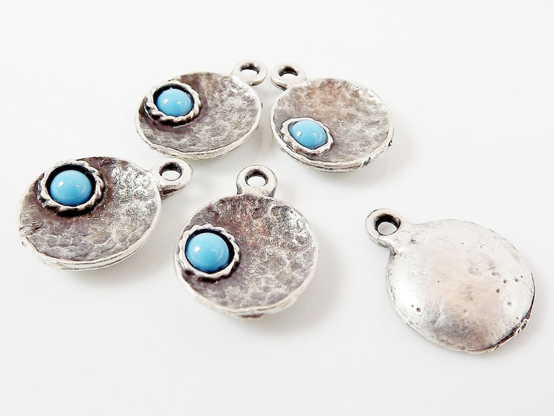 May include: Five silver charm pendants with a turquoise blue stone in the center. The pendants are round and have a textured surface.
