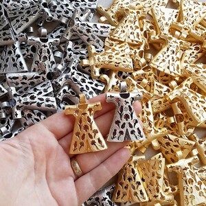 Pu&ograve; includere: Primo piano di ciondoli in metallo color oro e argento. I ciondoli hanno la forma di un vestito con intricati ritagli floreali. I ciondoli sono tenuti in mano, con un mucchio di ciondoli simili sullo sfondo.