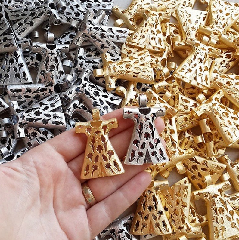 Pu&ograve; includere: Primo piano di ciondoli in metallo color oro e argento. I ciondoli hanno la forma di un vestito con intricati ritagli floreali. I ciondoli sono tenuti in mano, con un mucchio di ciondoli simili sullo sfondo.