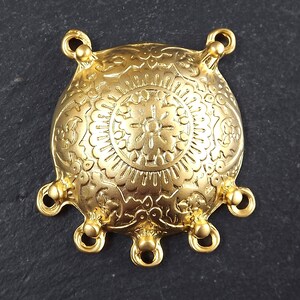May include: Gold-toned metal pendant with a round, ornate design and five loops. The pendant has a detailed floral pattern and a central sunburst motif.