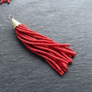 May include: A red beaded tassel with a gold metal top. The tassel is made of small, round beads that are strung together to create a long, flowing fringe.