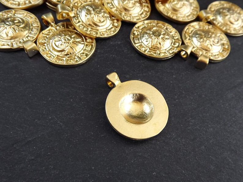 Pode incluir: Um grupo de pingentes de metal dourado com um design circular. Os pingentes t&ecirc;m um centro elevado e uma superf&iacute;cie texturizada. Um pingente est&aacute; deitado de costas, mostrando o interior liso e c&ocirc;ncavo.