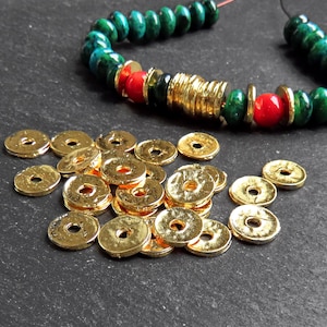 May include: A pile of gold-coloured metal beads with a hole in the centre. The beads are scattered on a dark grey surface. A green and red beaded bracelet is visible in the background.