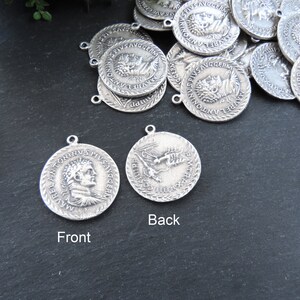May include: Two silver-toned metal pendants with a Roman emperor's profile on the front and a bird design on the back. The pendants have a rope-like border and are on a dark gray surface.