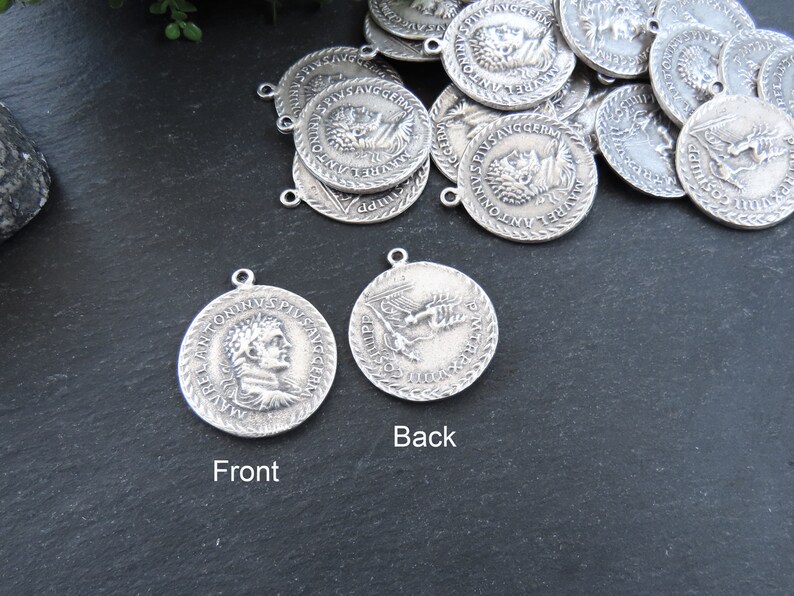 May include: Two silver-toned metal pendants with a Roman emperor's profile on the front and a bird design on the back. The pendants have a rope-like border and are on a dark gray surface.