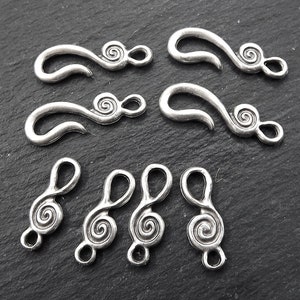 May include: A selection of silver-coloured metal jewellery clasps and charms. The clasps have a curved hook shape with a spiral design. The charms are shaped like treble clefs with a spiral design.