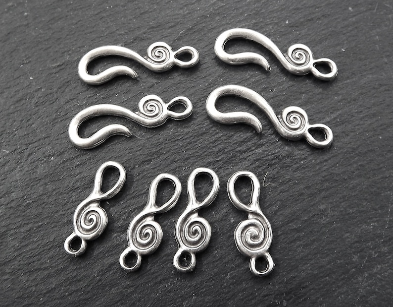 May include: A selection of silver-coloured metal jewellery clasps and charms. The clasps have a curved hook shape with a spiral design. The charms are shaped like treble clefs with a spiral design.